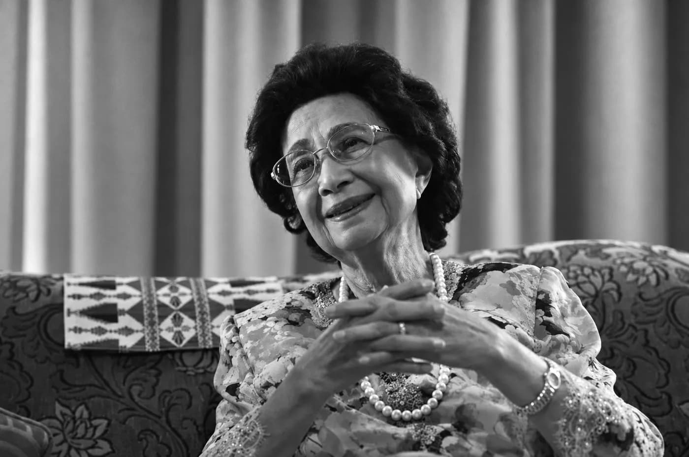 Black and white portrait of Tun Dr Hasmah Mohamad Ali, seated and smiling, hands clasped, wearing a pearl necklace.